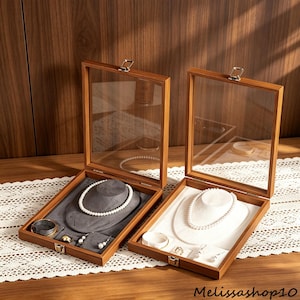 May include: Two open wooden jewelry display boxes with glass lids. One box features a gray velvet interior with a pearl necklace, earrings, and a bracelet. The other box has a white interior with a pearl necklace, earrings, and a bracelet.