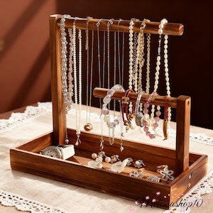 May include: A wooden jewelry organizer with a tray base and two horizontal bars for hanging necklaces and bracelets. The organizer displays various necklaces, bracelets, rings, and a watch. The wood has a warm, brown tone, and the jewelry includes pearls and gemstones.