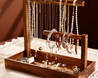 Op de afbeelding: Een houten sieradenorganizer met een dienbladbasis en twee horizontale staven om kettingen en armbanden op te hangen. De organizer toont diverse kettingen, armbanden, ringen en een horloge. Het hout heeft een warme, bruine tint en de sieraden bevatten parels en edelstenen.