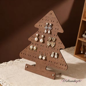 Può includere: Un espositore per orecchini in legno marrone a forma di albero di Natale. L'espositore ha fori per appendere gli orecchini. Diverse paia di orecchini, tra cui modelli con perle e cristalli, sono esposti.