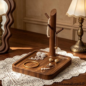 Könnte beinhalten: Ein Schmuck-Organizer aus dunklem Holz mit einem baumartigen Design. Er verfügt über eine Tablettbasis und einen vertikalen Pfosten mit Ästen zum Aufhängen von Halsketten und zum Präsentieren von Ohrringen. Verschiedene Schmuckstücke sind auf dem Tablett angeordnet.
