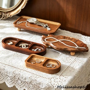 May include: A collection of wooden jewelry trays in various shapes and sizes. One rectangular tray holds a watch and necklace. Other trays display earrings, rings, and a pearl necklace. The trays are made of dark wood and sit on small wooden feet.