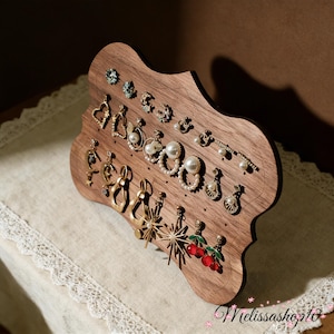 Può includere: Un espositore di orecchini in legno dalla forma unica, che mostra vari orecchini. Gli orecchini includono borchie, cerchi e pendenti, con perle, cristalli e altri elementi decorativi. L'espositore è in legno scuro e si trova su un panno bianco.