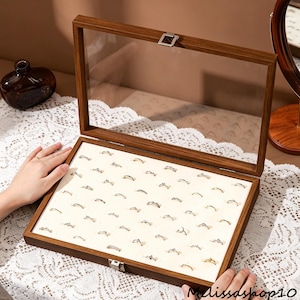 May include: A rectangular jewelry display case with a dark brown wooden frame and a clear glass lid. The interior features a cream-colored velvet insert with multiple slots designed to hold rings in an organized grid pattern. A metal latch secures the lid.