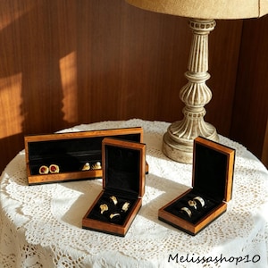Puede incluir: Tres joyeros de madera con exteriores de veta de madera marrón oscuro e interiores de terciopelo negro. Una caja rectangular larga contiene pendientes, mientras que dos cajas cuadradas más pequeñas contienen anillos dorados. El texto Melissashop10 es visible.