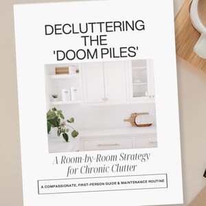 May include: A 5-page guide titled Decluttering Room by Room and Decluttering the Doom Piles. The cover features a photograph of a white kitchen with cabinets, a plant, and a wooden cutting board, with the subtitle A Room-by-Room Strategy for Chronic Clutter.