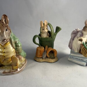 May include: Three ceramic figurines featuring animal characters. One is a rabbit in a green jacket, another is a rabbit in a watering can, and the third is a mouse with a broom. The figurines are painted in muted colors.