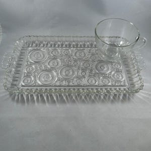 May include: A clear glass serving tray with a matching teacup. The rectangular tray features a textured surface with raised circles and a beaded edge. The teacup has a handle and sits on the tray. The set is designed for serving tea or small snacks.