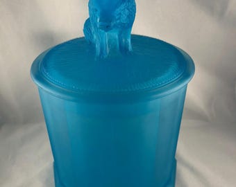 May include: A turquoise-colored glass jar with a lid featuring a sculpted bear figurine. The jar has an octagonal base and a textured lid. The bear is sitting on top of the lid. The jar is made of glass.