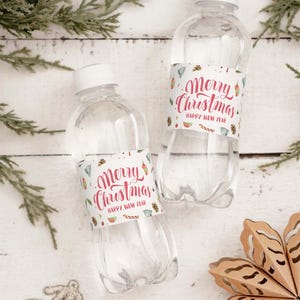 Könnte beinhalten: Zwei transparente Plastikwasserflaschen mit weißen Deckeln, jede mit einem Etikett mit der Aufschrift "Merry Christmas Happy New Year" in roter Schrift. Die Etiketten zeigen festliche Illustrationen. Auf einer weißen Holzoberfläche.