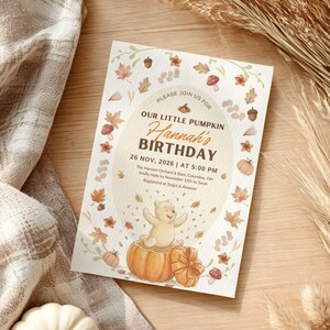 Könnte beinhalten: Eine rechteckige Geburtstagseinladungskarte mit einem zentralen Oval, das einen Cartoon-Bären in einem Kürbis zeigt. Der Text lautet: PLEASE JOIN US FOR OUR LITTLE PUMPKIN Hannah's BIRTHDAY 26 NOV, 2026 | AT 5:00 PM. Umrandet von Herbstblättern, Eicheln und Pilzen.