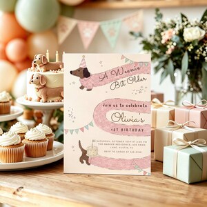 Op de afbeelding: Een rechthoekige verjaardagsuitnodiging met een illustratie van een lange teckel in een roze trui, feestelijke slingers en de tekst A Weenie Bit Older, Olivia's 1st Birthday, Saturday, October 10th at 2:00 PM, The Barker Residence, 456 Paws Lane, Austin, TX.
