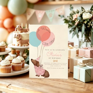 Op de afbeelding: Een rechthoekige verjaardagsuitnodiging met een bruine teckel in een roze trui en feesthoedje, die drie ballonnen vasthoudt. De tekst luidt A Weenie Bit Older, Join us to celebrate Olivia's 1st Birthday, inclusief de details van het feest.