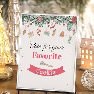 May include: A framed sign with the text "Vote for your Favorite Cookie" in pink and black. The sign is decorated with Christmas-themed illustrations and a banner with a whisk. The frame is silver.