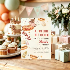 Op de afbeelding: Een rechthoekige verjaardagsuitnodiging met aquarelillustraties van teckels, hotdogs en feestartikelen. De tekst luidt: Olivia's A Weenie Bit Older, Join Us To Celebrate Olivia's 1st Birthday, zaterdag 10 oktober om 14:00 uur.