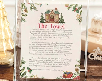 May include: A framed print with the text "The Towel" and a Christmas theme. The print features a house illustration, pine branches, and festive decorations. The text describes the towel's significance in everyday life and during the Christmas season.