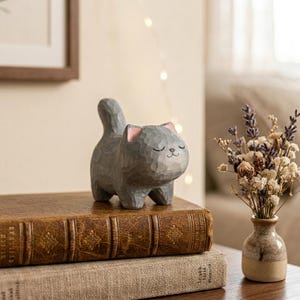 May include: A gray wooden cat figurine with pink accents on the ears, standing on a stack of vintage books. The cat has closed eyes and a smiling expression. A small vase of dried flowers sits nearby.