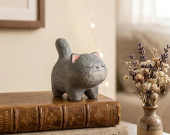 May include: A gray wooden cat figurine with pink accents on the ears, standing on a stack of vintage books. The cat has closed eyes and a smiling expression. A small vase of dried flowers sits nearby.