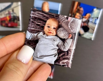 May include: A square photo magnet featuring a high-resolution color image of a baby wearing a light gray long-sleeved shirt with a small bear graphic, lying on a dark textured surface next to a soft gray plush toy, with a glossy finish.