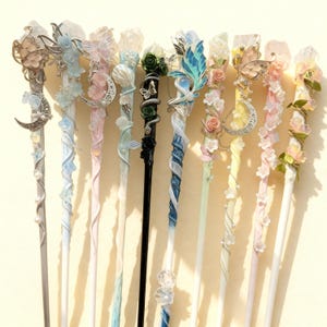 Puede incluir: Una colección de palillos decorativos para el cabello con diversos diseños. Cada palillo presenta un adorno diferente, incluyendo mariposas, flores y cristales. La paleta de colores incluye plata, rosa, azul y blanco, creando una estética etérea. Miden unos 20 cm.