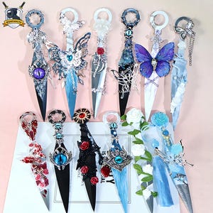 May include: An assortment of decorative daggers with detailed handles and blades. The daggers showcase butterfly, floral, and gemstone designs. Blades are silver and black, with blue, red, and purple accents. The handles are embellished with intricate metalwork.