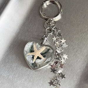 May include: A silver-toned keychain featuring a heart-shaped pendant filled with a small beige starfish and white sand. The pendant is attached to a circular ring and a dangling chain adorned with multiple small, flat, silver-colored star charms.