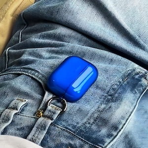 May include: A glossy, vibrant royal blue protective case for wireless earbuds, featuring a smooth, rounded rectangular shape and a black metal carabiner clip attached to a small loop, secured to the belt loop of light blue denim jeans.