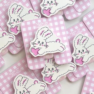 May include: A collection of white rabbit pins with pink heart accents, each attached to a light pink and white gingham display card featuring the text Made by LuckyStars. The rabbits have long ears, pink cheeks, and are holding a pink heart.