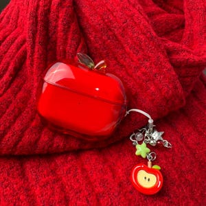 May include: A glossy, translucent red apple-shaped protective case for wireless earbuds. The case features a small green leaf accent on top and a dangling charm attachment with silver-toned metal links, a light green star, and a small red apple pendant.