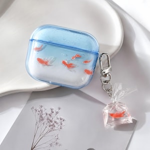 May include: A clear, light blue protective case for wireless earbuds featuring a gradient design with small orange goldfish illustrations. A silver metal keychain is attached to the side, holding a miniature clear plastic bag charm containing a goldfish.