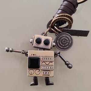 May include: A robot-shaped keychain with a bronze finish. The robot has black eyes and a black square on its chest, with decorative details. It is attached to a brown leather loop and metal rings.