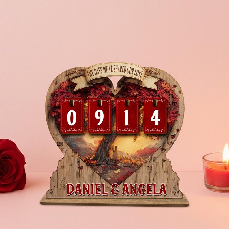 Puede incluir: Calendario de cuenta regresiva de madera en forma de coraz&oacute;n con el texto "THE DAYS WE'VE SHARED OUR LOVE" en una pancarta. El calendario muestra "0914" y los nombres "DANIEL & ANGELA". El fondo presenta un &aacute;rbol y una pareja.