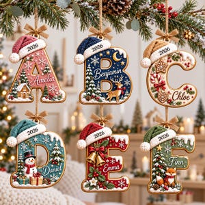 May include: Six hanging Christmas ornaments shaped as letters A, B, C, D, E, and F. Each letter features a festive winter scene, a personalized name, and a miniature Santa hat with the year 2026 printed on the brim, suspended by twine bows.