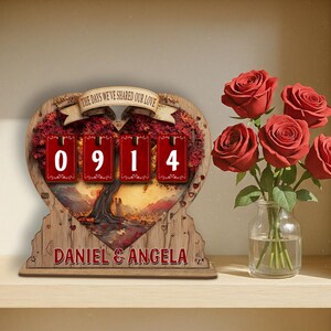 Puede incluir: Un calendario de cuenta regresiva de madera en forma de coraz&oacute;n con el texto "THE DAYS WE'VE SHARED OUR LOVE" y los n&uacute;meros "0914". La parte inferior del calendario dice "DANIEL & ANGELA". Un jarr&oacute;n de rosas rojas est&aacute; a la derecha.