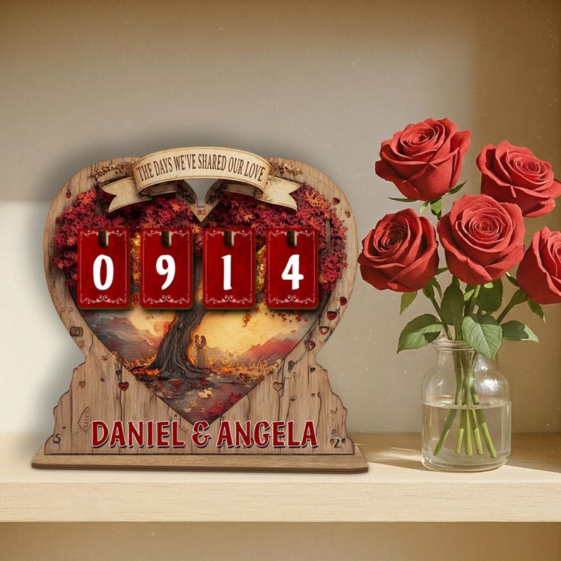 Puede incluir: Un calendario de cuenta regresiva de madera en forma de coraz&oacute;n con el texto "THE DAYS WE'VE SHARED OUR LOVE" y los n&uacute;meros "0914". La parte inferior del calendario dice "DANIEL & ANGELA". Un jarr&oacute;n de rosas rojas est&aacute; a la derecha.