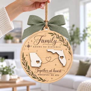 Op de afbeelding: Een rond houten ornament met een saliegroene stoffen strik en een touwtje om op te hangen. Het oppervlak toont de tekst "Family KNOWS NO DISTANCE" en "Together at heart wherever we are" met lasergesneden kaarten van Georgia en Florida verbonden door een stippellijn en hartjes.