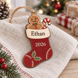 Peut inclure: Une décoration de Noël en bois en forme de chaussette rouge avec un revers clair. La chaussette porte le prénom Ethan, l'année 2026 et un brin de houx vert. Un bonhomme en pain d'épices marron et un sucre d'orge rouge et blanc dépassent.