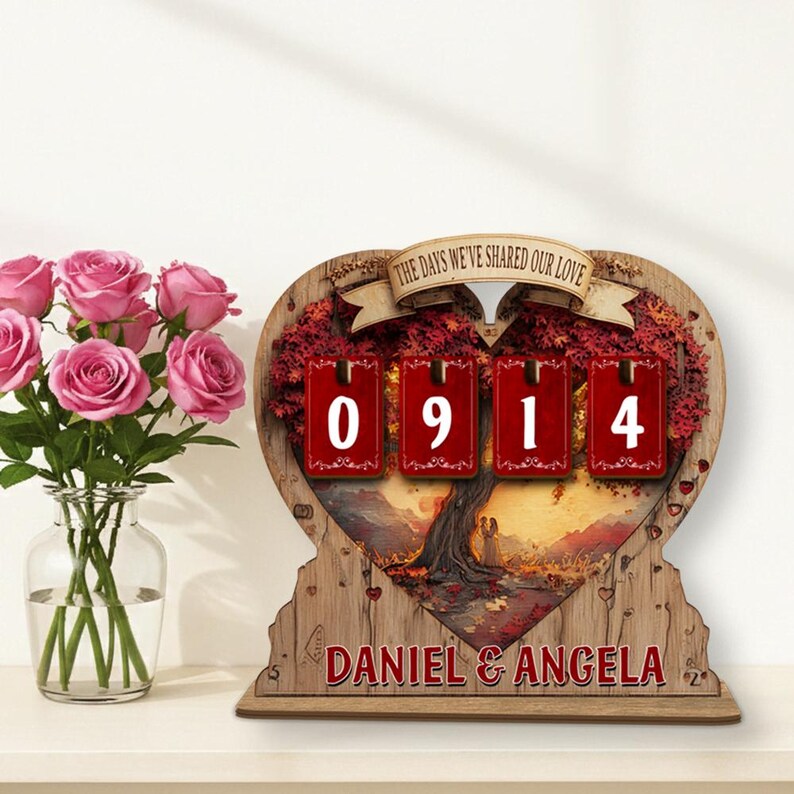 Puede incluir: Calendario de cuenta regresiva de madera en forma de coraz&oacute;n con el texto "THE DAYS WE'VE SHARED OUR LOVE". El calendario muestra "0914" en paneles rojos. La base dice "DANIEL & ANGELA". Un jarr&oacute;n de rosas rosas est&aacute; a la izquierda.