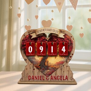 Puede incluir: Adorno de madera en forma de coraz&oacute;n con el texto "THE DAYS WE'VE SHARED OUR LOVE" y los nombres "DANIEL & ANGELA". Presenta un dise&ntilde;o de &aacute;rbol con los n&uacute;meros "0914" en tarjetas rojas. Corazones decorativos cuelgan en el fondo.