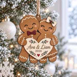 May include: A wooden gingerbread couple ornament featuring a brown finish, white icing details, and a maroon bow tie. The pair holds a cream-colored heart inscribed with the text Ava & Liam 2026, decorated with green leaves and a small red heart.