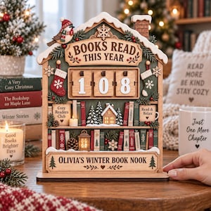 May include: A wooden house-shaped book tracker with a flip-number display showing 108, shelves with miniature books, a bird, stockings, and text reading BOOKS READ THIS YEAR and OLIVIA'S WINTER BOOK NOOK, featuring integrated warm lighting.