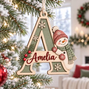 May include: A wooden letter A Christmas ornament featuring a sage green finish, the name Amelia in red script, a smiling snowman wearing a red hat and green scarf, small white snowflakes, and a cluster of green pine needles with red berries.