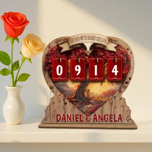 Puede incluir: Un calendario perpetuo de madera en forma de coraz&oacute;n con el texto "THE DAYS WE'VE SHARED OUR LOVE" y los nombres "DANIEL & ANGELA". El calendario muestra los n&uacute;meros "0914" en tarjetas rojas. A la izquierda, un jarr&oacute;n con una rosa roja y amarilla.