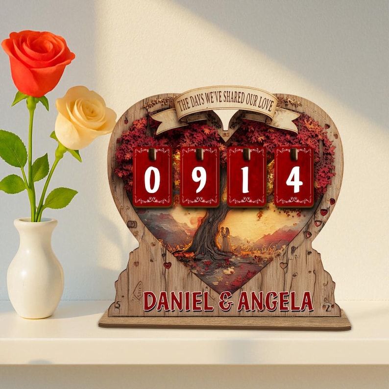 Puede incluir: Un calendario perpetuo de madera en forma de coraz&oacute;n con el texto "THE DAYS WE'VE SHARED OUR LOVE" y los nombres "DANIEL & ANGELA". El calendario muestra los n&uacute;meros "0914" en tarjetas rojas. A la izquierda, un jarr&oacute;n con una rosa roja y amarilla.