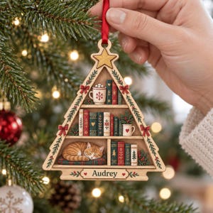 May include: A triangular wooden Christmas tree ornament featuring shelves with miniature books, a mug with a snowflake, a mug with a heart, and a sleeping orange tabby cat on a cushion. The base is engraved with the name Audrey and a red ribbon.