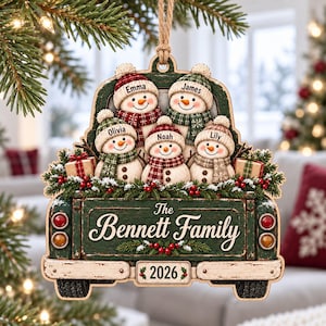 May include: A wooden Christmas ornament shaped like a truck bed carrying five snowmen wearing hats and scarves labeled Emma, James, Olivia, Noah, and Lily. The truck features the text The Bennett Family and 2026, decorated with pine and red berries.