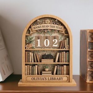 May include: Wooden bookshelf ornament with the text "BOOKS READ THIS YEAR" and "102". The shelves are filled with miniature books, a black cat figurine, and the text "OLIVIA'S LIBRARY".