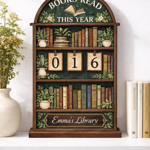 Może przedstawiać: Drewniany regał na książki z napisem "BOOKS READ THIS YEAR" na górze. Regał ma trzy półki wypełnione książkami, roślinami i liczbami "016". Na dole regału napis "Emma's Library".