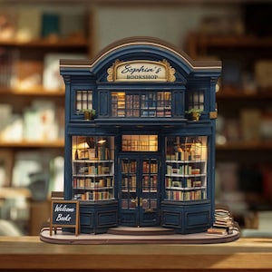 Könnte beinhalten: Ein detailliertes Miniaturmodell eines blauen Buchladens mit der Aufschrift "Sophia's BOOKSHOP" über dem Eingang. Die beleuchteten Fenster zeigen Bücherregale. Ein kleines Tafelschild zeigt "Welcome Books".
