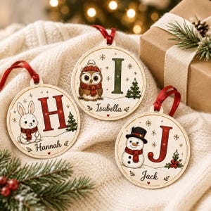 May include: Three round wooden Christmas ornaments with red ribbon hangers. One features a rabbit and the letter H with the name Hannah. Another shows an owl and the letter I with the name Isabella. The third displays a snowman and the letter J with the name Jack.